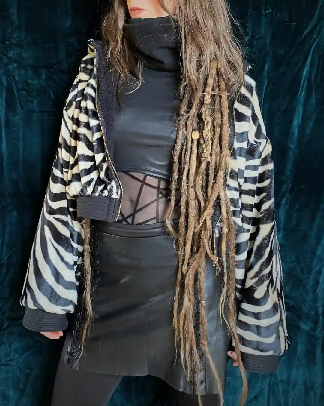 Frontansicht einer Trägerin mit langer Dreadlockfrisur, die eine Zebra-Felljacke über einem eng anliegenden schwarzen Top mit Mesh-Elementen und einem geschnürten Lederrock trägt. Der Style wirkt edgy und alternativ. Die Felljacke ist geöffnet und zeigt das warme Innenfellfutter.