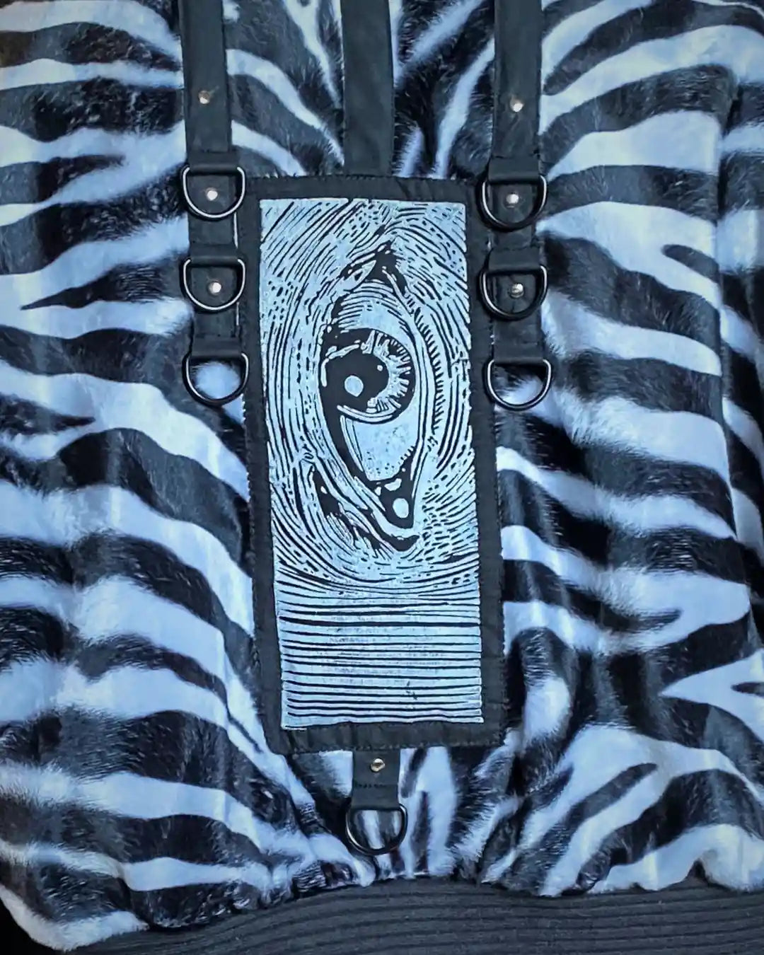 Handgefertigte Zebrafell-Jacke aus veganem Kunstfell mit Kapuze und schwarzem Patch mit Auge-Print und D-Ring Applikationen am Rücken – Slow Fashion Darkwear von IUVA CRAFT STUDIO